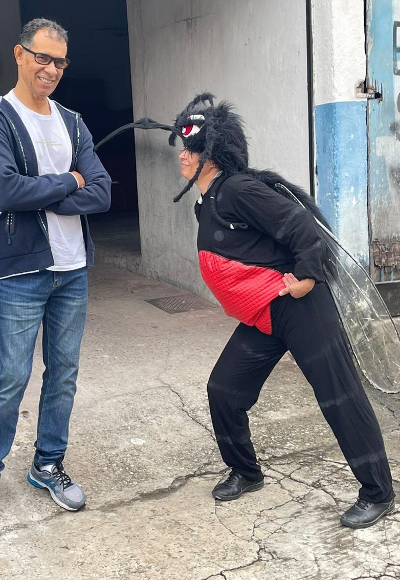 A foto mostra uma agente de saúde fantasiada de mosquito da dengue; ela está na rua, e interage com um homem, que sorri